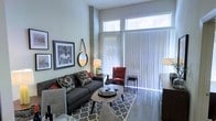 This thoughtfully designed single-floor space makes a striking first impression with its soaring ceilings and dramatic floor-to-ceiling windows that flood the interior with abundant natural light. The living area showcases a refined industrial aesthetic, highlighted by an exposed concrete column that adds an urban, loft-like character to the space.

The living room is generously proportioned and styled for both comfort and sophistication. A charcoal gray sofa anchors the seating area, complemented by a bold red accent chair that adds a pop of color. A geometric-patterned area rug defines the space, while a sleek oval coffee table serves as a central gathering point. Curated wall art and a round decorative mirror contribute to the room's polished, contemporary feel.

Large vertical blinds along the expansive windows offer flexible light control while maintaining the airy, open quality of the space. Polished concrete flooring runs throughout, reinforcing the modern industrial tone while remaining practical and durable. The high clerestory windows further amplify the sense of vertical space and openness.

Overall, the space blends urban edge with warm, inviting touches, creating an environment that feels both stylish and livable — ideal for those who appreciate contemporary design with character.