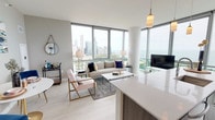 This contemporary high-rise residence offers a thoughtfully designed open-concept layout with light wood flooring and a neutral color palette that creates a bright, cohesive atmosphere throughout. A welcoming entryway with modern wall décor leads into the main living area, where expansive floor-to-ceiling windows frame breathtaking panoramic views of the Chicago skyline, Grant Park, and Lake Michigan. The living room is bathed in natural light and features a corner window configuration that maximizes the sweeping vistas on multiple sides. The kitchen is equipped with stainless steel appliances, white cabinetry accented by warm wood uppers, a full-height tile backsplash, and a generous waterfall-edge quartz island with bar seating — ideal for both cooking and entertaining. A dedicated in-unit stacked washer and dryer adds everyday convenience. The bedroom is highlighted by a bold navy accent wall, floor-to-ceiling windows continuing the skyline views, and a well-organized closet with built-in shelving. The bathroom features large-format tile flooring, a wood-toned vanity with ample storage, and a clean, contemporary design. Throughout the residence, the interplay of modern finishes, abundant natural light, and dramatic urban views creates a refined and functional living environment.