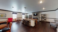 Located at 2601–2607 Lillian Ln in Beavercreek, Ohio, this single-floor communal gathering space is thoughtfully designed to accommodate a variety of social settings. The room is anchored by a striking floor-to-ceiling stacked stone fireplace with a wall-mounted flat-screen television above it, creating a natural focal point. Adjacent to the fireplace is a stone-accented bar-height counter with a granite-style surface and upholstered bar stools, ideal for casual conversation or informal gatherings.

Dark hardwood flooring extends throughout the entire space, complemented by contrasting dark crown molding that frames the neutral-toned walls. Recessed lighting and pendant lights provide consistent illumination across the room. Multiple seating configurations are distributed throughout, including round dining tables with wooden chairs, vibrant red tufted armchairs paired with low round coffee tables, and gray upholstered lounge chairs arranged for quiet conversation. Framed artwork adorns the walls, adding visual interest and a welcoming ambiance.

A secondary stone-clad reception or service counter near the entrance features pendant lighting overhead. Windows fitted with dark curtains allow natural light to filter in. The overall layout offers a versatile, well-appointed environment suitable for both lively social events and relaxed everyday use.