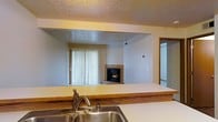This single-floor space is designed with an open-concept layout that connects the kitchen and living room, creating a seamless flow between the two areas. The kitchen features a light-colored countertop with warm wood-tone edging and a stainless steel sink, positioned at a pass-through opening that overlooks the living area. This design element enhances the sense of openness while maintaining a functional separation between spaces.

The living room serves as the focal point of the unit, highlighted by a tile-surround fireplace that adds warmth and character to the space. A sliding glass door with vertical blinds allows natural light to filter into the room and provides potential access to an outdoor area. The neutral wall tones throughout create a clean, versatile backdrop suitable for a variety of interior styles.

A textured popcorn ceiling is present throughout the space, consistent with the overall construction aesthetic. A doorway visible from the kitchen pass-through suggests additional rooms or a hallway beyond. The layout offers a practical and connected living environment, ideal for everyday comfort and functionality.