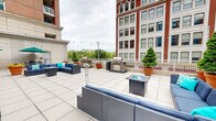Located at 100 Seventh St in the heart of downtown Pittsburgh, this rooftop terrace offers a generously sized outdoor amenity space designed for both relaxation and entertainment. The terrace is finished with large-format light-colored tile pavers that extend across its L-shaped footprint, creating a clean, urban aesthetic throughout.

Multiple distinct seating zones are arranged across the terrace, each furnished with wicker sectional sofas and chairs in navy blue cushions, centered around fire pit tables — ideal for gathering during cooler evenings. Additional lounging options include classic wrought-iron chaise lounges with blue cushions, perfectly positioned for open-air relaxation. Teal market umbrellas provide shaded seating areas with bistro-style table and chair sets nearby.

Dedicated grilling stations with built-in outdoor grills offer a functional cooking area, making the space well-suited for social events and cookouts. Potted topiaries, ornamental shrubs, and terracotta planters add greenery and a polished, curated feel throughout the space.

String lights are strung overhead across multiple sections of the terrace, lending a warm ambiance for evening use. The terrace is framed by views of Pittsburgh's surrounding downtown skyline and neighboring historic brick architecture, with glass-door access points connecting directly to the interior. The overall atmosphere is vibrant, social, and thoroughly urban.