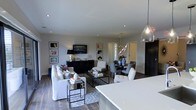 This well-appointed single-floor residence showcases a thoughtfully designed open-concept layout that fluidly connects the kitchen, dining, and living areas into one cohesive and inviting environment. The living area is anchored by a plush white sofa set arranged around a sleek black metal coffee table, all set atop a patterned area rug over rich dark hardwood flooring. A wall-mounted flat-screen television is flanked by a series of framed artworks and a dark credenza, adding a curated, gallery-like quality to the space. The dining area features a round table surrounded by upholstered chairs, illuminated by a sculptural brass branching chandelier that serves as a striking focal point. Adjacent to the dining space, a doorway framed in crisp white millwork leads further into the home, offering a sense of depth and continuity. The kitchen is defined by a generous white countertop island and a pendant glass light fixture, blending modern functionality with refined aesthetics. Recessed lighting throughout ensures the entire space is evenly and warmly lit. The neutral color palette of whites, grays, and dark accents creates a sophisticated, timeless ambiance suitable for both everyday living and entertaining.