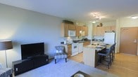 This single-floor space offers an open-concept layout that connects the kitchen and living area, creating a cohesive and functional environment. The kitchen is well-appointed with light wood cabinetry that extends to the upper walls, providing generous storage throughout. White appliances, including a range, microwave, and refrigerator, complement the neutral color palette, while a dark-toned countertop adds a grounding contrast. A peninsula with a granite-style countertop and bar seating serves as a natural divider between the kitchen and the dining area, making it ideal for casual dining and entertaining. The living area features soft, light-toned carpeting that contrasts warmly with the hardwood flooring in the kitchen, helping to define each zone within the open plan. Neutral grey walls keep the space feeling bright and versatile, while track lighting in the kitchen ensures the workspace is well-lit. The overall ambiance is clean, welcoming, and practical — well-suited for comfortable everyday living.