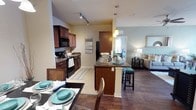 This well-appointed 2-bedroom, 2-bathroom residence offers a thoughtfully designed single-floor layout that maximizes comfort and functionality. The open-concept floor plan seamlessly connects the kitchen, dining, and living areas, creating a cohesive and inviting atmosphere. The kitchen is a standout feature, showcasing rich dark wood cabinetry, dark granite-style countertops, a tile backsplash, and black appliances including a range, microwave, and refrigerator. A central kitchen island with seating provides additional prep space and serves as a natural gathering point. Track lighting and pendant lights illuminate the space with a warm, functional glow. Hardwood-style flooring flows through the main living areas, complemented by tile in the kitchen, adding both style and practicality. The dining area accommodates a full table setting, ideal for entertaining. The living room is bright and open, offering a comfortable retreat. A notable highlight of this residence is the solarium, adding a unique, light-filled bonus space not commonly found in similar layouts. The two bedrooms and two full bathrooms provide ample accommodation, while the overall neutral color palette with teal accents contributes to a calm, modern ambiance throughout.