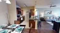 This well-appointed 2-bedroom, 2-bathroom residence offers a thoughtfully designed single-floor layout that maximizes comfort and functionality. The open-concept floor plan seamlessly connects the kitchen, dining, and living areas, creating a cohesive and inviting atmosphere. The kitchen is a standout feature, showcasing rich dark wood cabinetry, dark granite-style countertops, a tile backsplash, and black appliances including a range, microwave, and refrigerator. A central kitchen island with seating provides additional prep space and serves as a natural gathering point. Track lighting and pendant lights illuminate the space with a warm, functional glow. Hardwood-style flooring flows through the main living areas, complemented by tile in the kitchen, adding both style and practicality. The dining area accommodates a full table setting, ideal for entertaining. The living room is bright and open, offering a comfortable retreat. A notable highlight of this residence is the solarium, adding a unique, light-filled bonus space not commonly found in similar layouts. The two bedrooms and two full bathrooms provide ample accommodation, while the overall neutral color palette with teal accents contributes to a calm, modern ambiance throughout.