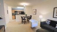 This single-floor residence showcases an open-concept floor plan that integrates the living, dining, and kitchen areas into one cohesive, functional space. The layout promotes a natural flow between zones, making the interior feel spacious and inviting.

The living area features neutral-toned carpeting that extends throughout the main space, contributing to a warm and comfortable atmosphere. Modern seating, including a white upholstered chair and a dark sofa, is arranged to complement the room's open feel. A table lamp adds ambient lighting to the space.

Adjacent to the living area, a compact dining nook accommodates a dark bistro-style table with matching chairs, ideal for everyday meals. A framed botanical print on the wall adds a subtle decorative touch.

The kitchen is defined by light wood cabinetry, a breakfast bar peninsula, and black appliances, including a refrigerator and range. The peninsula serves as a visual divider between the kitchen and the dining area while maintaining the open feel of the layout. Overhead fluorescent lighting illuminates the kitchen workspace effectively.

Overall, the space presents a clean, neutral aesthetic with practical design choices suited for comfortable, everyday living.