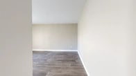 Located at 7000 Red Fox Trail in Shreveport, Louisiana, this single-floor unit offers a functional and efficiently organized layout comprising a living room, kitchen, hallway, and bathroom. Throughout the space, durable wood-look vinyl plank flooring in a warm gray tone provides a cohesive and modern aesthetic, complemented by crisp white baseboards and neutral taupe walls that create a clean, inviting atmosphere. The living room is the largest area of the unit, featuring vaulted ceilings that lend a sense of openness beyond its compact footprint. A sliding glass door floods the room with natural light and provides an outdoor view, enhancing the airy feel of the space. The kitchen is thoughtfully equipped with white shaker-style cabinetry, granite-look countertops, and black appliances including an electric range with overhead ventilation and a full-size refrigerator. A peninsula-style counter divides the kitchen from the main living area, adding both functionality and definition. The bathroom features a full tub-shower combination with white surround panels and chrome fixtures, maintaining a clean and practical finish. A hallway connects the rooms and includes a closet with a white panel door. Overall, the unit presents a neat, move-in ready condition with a consistent, contemporary style.