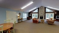 This single-story space is designed to feel open and inviting, anchored by dramatic vaulted ceilings that rise to a striking peak along one wall, clad in dark brown paneling. A large triangular window at the apex of the vaulted section floods the room with natural light and frames views of surrounding greenery, complemented by additional windows fitted with blinds along the side walls and glass-paneled double doors leading to the outdoors. The centerpiece of the space is a floor-to-ceiling stone fireplace with a classic mantle, lending warmth and a timeless architectural character to the room. The interior is finished with light blue walls and wall-to-wall carpeting in a neutral tone, contributing to a calm, comfortable atmosphere. Seating includes a plush sofa and a recliner chair, arranged to encourage relaxed conversation or quiet enjoyment. A wooden console table, decorative wall clock, and framed artwork add homey, personal touches throughout. Overhead fluorescent lighting ensures the space remains bright and well-lit at all times. The overall layout is spacious and accessible, with a single-floor design that promotes ease of movement throughout the area.