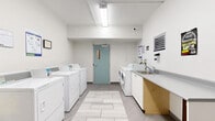 This shared laundry facility is thoughtfully designed to accommodate multiple users simultaneously. The space is bright and clean, with white walls and large-format tile flooring that runs throughout, lending a modern, orderly aesthetic. Overhead fluorescent lighting ensures the room is well-lit at all times.

The laundry area is lined with a row of top-loading washing machines and dryers along one wall, complemented by front-loading washer and dryer units on the opposite side, offering users a variety of machine options. A utility sink with a grey countertop provides a practical station for hand-washing or pre-treating garments.

A dedicated folding and seating area features an extended grey countertop with white cabinetry below and wooden barstools, providing a comfortable surface for sorting and folding laundry. A large wall-mounted mirror and framed artwork add a welcoming touch to this corner of the room.

Additional amenities include wall-mounted informational panels, a bulletin board near the entrance, and a security camera for safety. A teal-painted door with a small window serves as the primary access point. The overall layout is spacious and functional, designed to make laundry tasks as efficient and convenient as possible.