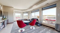 Located on the fifth floor at 839 16th St SW in Rochester, Minnesota, this contemporary lounge offers a well-appointed communal gathering space with a thoughtful, modern design. The open-plan layout is anchored by an expansive bank of large windows that wrap around the space, flooding the interior with abundant natural light and offering sweeping elevated views of the surrounding Rochester landscape, including treetops and neighboring rooftops.

The main living area is furnished with a plush charcoal sofa, colorful accent chairs, and a sleek round coffee table, creating an inviting environment for relaxation or social interaction. A pair of bold red swivel chairs on polished chrome bases serve as striking focal points throughout the space. Patterned carpet in muted gray tones adds texture underfoot while complementing the neutral warm-beige walls.

A dedicated dining nook features high-top tables paired with red bar-height chairs, with direct access to an outdoor terrace, extending the usable area and enhancing the connection to the outdoors. The built-in kitchenette along one wall includes cabinetry, counter space, and appliances, supporting everyday convenience. Recessed ceiling lighting and decorative framed artwork contribute to a polished, community-oriented ambiance. Restroom access and elevator entry points are also conveniently located within the space.