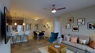 This single-floor residence offers a well-designed open-concept layout that seamlessly connects the living, dining, and kitchen areas into one cohesive and functional space. The living area is anchored by warm wood-tone flooring that extends throughout, creating a consistent and inviting atmosphere. A ceiling fan with integrated lighting sits overhead, ensuring both comfort and ambient illumination. The living space is furnished with a plush sofa, a rustic wooden coffee table, and a striking teal accent chair, adding a pop of color and personality to the neutral palette. Framed artwork is thoughtfully arranged along the walls, contributing to an elevated aesthetic. The dining area features a compact table with dark chairs, accompanied by a pendant light fixture above, making it a cozy spot for meals. Adjacent to the dining space, the kitchen is visible with light cabinetry and a counter-height seating area, maintaining the open and airy feel of the floor plan. Neutral-toned walls throughout keep the space feeling bright and versatile. The layout is efficient yet spacious, with interior doors providing access to additional rooms, making this a comfortable and well-appointed single-level home.