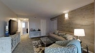 This single-floor residence offers a comfortable and thoughtfully arranged living environment. The centerpiece of the home is the living room, which immediately draws attention with its striking stone accent wall, finished in warm, earthy tones that add texture and visual depth to the space. A wall-mounted sconce highlights the stonework, casting a warm ambient glow throughout the room. The living area is carpeted in a neutral tone, complementing the soft gray walls and creating a cozy, welcoming atmosphere. Furnished with a light taupe sofa accented with decorative throw pillows and a patterned area rug beneath a sleek glass-top coffee table, the space balances style with everyday comfort. A console table along one wall provides additional display and storage space. Bifold and paneled doors offer access to closet storage, keeping the layout organized and practical. A hallway connects the living room to the rest of the home, where additional rooms extend the functional footprint of the space. Natural light filters through the adjoining areas, enhancing the overall brightness of the interior. The cohesive design palette of neutral tones and warm accents throughout creates a harmonious and inviting living environment.