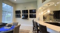 This single-floor entertainment space is thoughtfully designed for hosting and leisure, combining modern amenities with a welcoming atmosphere. The focal point of the main area is a striking wall-mounted arrangement of four flat-screen televisions, ideal for watching multiple events simultaneously. A dedicated media shelf beneath the screens keeps equipment neatly organized. Light, neutral-toned walls and tile flooring contribute to the bright, clean aesthetic throughout. Adjacent to the viewing area, a curved bar counter with elegant white countertops and upholstered, nail-head-trimmed bar seating provides a sophisticated spot for guests to gather. Pendant lights with cylindrical glass shades hang above the bar, casting a warm glow over the space. The nearby kitchen area features dark cabinetry, a mosaic tile backsplash, and recessed ceiling details, adding depth and visual interest. A cozy seating arrangement with plush chairs and a patterned area rug anchors the lounge zone, creating a comfortable retreat for relaxed conversation. The open-concept layout encourages flow between the entertainment, bar, and kitchen areas, making this space well-suited for a variety of social occasions.