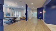 The Metro Clubhouse is a thoughtfully designed single-floor community gathering space that blends modern aesthetics with comfortable, functional living. The interior is defined by a striking color palette of crisp whites and bold navy blue, carried through bold architectural columns, accent walls, and a dramatic feature wall. That centerpiece wall showcases an intricate geometric, textured panel design finished in deep navy, housing a built-in fireplace and a mounted television — a focal point that anchors the main lounge area. Rich hardwood-style flooring flows throughout, complementing plush seating arrangements, a tufted sectional sofa, and curated greenery that adds warmth and life to the space. A dedicated game zone features an air hockey table beneath a geometric cage chandelier, set on decorative patterned tile flooring, framed by a bold navy entryway and brass sconce lighting. A dining and co-working area boasts floor-to-ceiling windows that flood the space with natural light and offer direct views of the outdoor courtyard pool area. A billiards table provides additional entertainment, while cozy seating nooks with mustard accent chairs offer inviting spots for conversation. Curated artwork, indoor plants in gold planters, and carefully chosen lighting fixtures throughout give the Metro Clubhouse a polished, upscale ambiance.