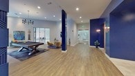 The Metro Clubhouse is a thoughtfully designed single-floor community gathering space that blends modern aesthetics with comfortable, functional living. The interior is defined by a striking color palette of crisp whites and bold navy blue, carried through bold architectural columns, accent walls, and a dramatic feature wall. That centerpiece wall showcases an intricate geometric, textured panel design finished in deep navy, housing a built-in fireplace and a mounted television — a focal point that anchors the main lounge area. Rich hardwood-style flooring flows throughout, complementing plush seating arrangements, a tufted sectional sofa, and curated greenery that adds warmth and life to the space. A dedicated game zone features an air hockey table beneath a geometric cage chandelier, set on decorative patterned tile flooring, framed by a bold navy entryway and brass sconce lighting. A dining and co-working area boasts floor-to-ceiling windows that flood the space with natural light and offer direct views of the outdoor courtyard pool area. A billiards table provides additional entertainment, while cozy seating nooks with mustard accent chairs offer inviting spots for conversation. Curated artwork, indoor plants in gold planters, and carefully chosen lighting fixtures throughout give the Metro Clubhouse a polished, upscale ambiance.