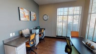 This single-floor business center is thoughtfully designed to accommodate both collaborative meetings and individual work needs. The space is divided into two primary functional areas that complement each other in style and purpose.

The conference area serves as the centerpiece of the space, anchored by a sleek, dark-toned conference table surrounded by a mix of vibrant green and orange ergonomic chairs. Soaring ceilings amplify the sense of openness, while an impressive arrangement of tall, floor-to-ceiling windows floods the room with abundant natural light and offers views of the surrounding outdoor greenery. A bold, colorful abstract artwork adorns the wall, adding a creative and dynamic energy to the environment. Wood-look flooring runs throughout, lending warmth and cohesion.

Adjacent to the conference area is a dedicated computer workstation nook, finished in cool gray tones. Built-in cabinetry with a wide countertop supports multiple computer workstations and a printer, making it fully equipped for productivity. Complementary abstract wall art and a wall-mounted clock complete this area's functional yet polished aesthetic.

Overall, the space strikes a balance between professional utility and modern design sensibility, making it an ideal environment for focused work, meetings, and collaborative sessions.