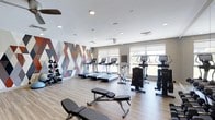 Located at 665–711 Commerce Dr in Fairfield, Connecticut, this fitness center offers a well-appointed, single-floor workout facility designed to support a range of exercise routines. The space is immediately distinguished by a striking geometric accent wall, featuring an eye-catching pattern of triangles in muted tones of terracotta, gray, and white, lending the room a contemporary and energizing aesthetic.

The cardio zone is equipped with multiple treadmills and elliptical machines, all positioned to face large windows fitted with roller shades that allow natural light to fill the space while maintaining comfort. A stationary bike adds additional variety to the cardio offerings. The open floor plan provides ample room to move freely between equipment.

A dedicated free weights area lines one wall, featuring a neatly organized dumbbell rack with a wide range of weights, alongside an adjustable workout bench positioned centrally for versatile strength training exercises. The light wood-toned flooring throughout the room creates a clean, modern look while remaining durable and easy to maintain.

Recessed ceiling lighting supplements the natural light from the windows, ensuring the space is bright and well-lit at any time of day. The neutral wall tones and uncluttered layout contribute to a focused, professional atmosphere well-suited for both casual and dedicated fitness routines.
