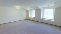 This single-floor space presents a generously sized open room finished with neutral wall-to-wall carpeting in a warm beige tone, creating a clean and versatile foundation. The room's most notable architectural feature is a subtle stepped or tray-like ceiling detail near the center, adding modest visual dimension to the otherwise smooth, white ceiling. A small chandelier with multiple light fixtures anchors one corner of the ceiling, providing an overhead lighting focal point.

Two windows bring in a notable amount of natural light, brightening the cream-colored walls and contributing to the room's open, airy feel. The windows feature low sills with ledges, which could serve as seating or display areas. White baseboard trim runs consistently along the perimeter of the room, offering a clean contrast against the light carpeting.

The walls are painted in a soft, neutral white-cream tone, maintaining a bright and understated aesthetic throughout. The overall ambiance of the space is calm and neutral, offering a blank canvas well-suited to a variety of uses and furnishing arrangements. The room feels open and uncluttered, with ample floor space to accommodate diverse configurations.