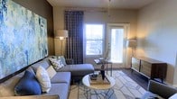 The living room serves as the centerpiece of this single-story space, designed with both comfort and style in mind. Hardwood-style flooring runs throughout, adding warmth and a sense of continuity. A large window dressed with patterned navy curtains allows an abundance of natural light to fill the room, while a glass-paneled door provides direct access to an outdoor area, further enhancing the bright, airy atmosphere.

The room features a generous sectional sofa in a neutral gray tone, complemented by an assortment of decorative throw pillows in coordinating blues and grays. A round coffee table anchors the seating area atop a geometric-patterned area rug, creating a well-defined and inviting gathering space. A large abstract canvas on the wall adds a bold artistic focal point, while a dresser and carefully placed floor lamps contribute functional yet stylish accents. The warm taupe wall tones tie the design elements together cohesively, resulting in a living space that is both welcoming and thoughtfully curated.