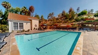 Located in Vista, CA, this well-appointed outdoor pool facility offers a resort-style experience under the Southern California sun. At its center is a generously sized rectangular lap pool with clearly marked lanes, ideal for both recreational swimming and fitness use. The surrounding paver deck provides ample space for relaxation, lined with numerous chaise lounge chairs and shaded by patio umbrellas positioned throughout the area.

Adjacent to the main pool is a circular spa, accessible via a short staircase and framed by a compact seating area with umbrella-shaded bistro tables and chairs — a comfortable spot to unwind. A pergola-covered zone on one side of the deck adds architectural interest and additional shaded seating, complemented by a built-in barbecue, making it well-suited for outdoor gatherings.

A pool house structure anchors one end of the facility, featuring a red clay tile roof consistent with a Spanish-influenced architectural style. It provides access to posted pool rules and utility amenities for the area. The entire space is framed by mature trees and vibrant landscaping, with a natural hillside rising in the background, adding privacy and a lush, scenic quality to the environment. Clear blue skies overhead enhance the bright, open-air ambiance of this inviting community amenity.