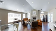 This single-story space showcases a thoughtfully designed living area centered around a striking stacked-stone fireplace, which serves as the room's focal point. The fireplace surround extends to the vaulted ceiling, creating a dramatic vertical element that draws the eye upward and enhances the sense of height and openness throughout the space.

The vaulted ceiling, fitted with recessed lighting, adds architectural interest while contributing to the airy, expansive feel of the room. Rich, dark hardwood flooring runs throughout, providing warmth and grounding the space with a classic, polished aesthetic.

Natural light is a defining feature, with multiple windows fitted with horizontal blinds lining the walls, including arched accent windows that add a refined architectural touch. The generous window placement ensures bright, well-lit interiors throughout the day.

The living area is arranged to encourage comfortable conversation, with cognac-toned leather seating complemented by patterned accent chairs around a central coffee table. A sleek console table positioned along one wall provides additional display and storage functionality, completing this well-appointed, cohesive living space.