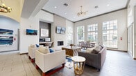 Located along Essex Lane in Houston's upscale 77027 corridor, this single-floor community amenity and leasing space is thoughtfully designed to offer both function and comfort. The entrance opens into a welcoming lobby featuring warm hardwood-style flooring, bold globe chandeliers, and a seating area with crisp white armchairs accented by royal blue pillows, set atop a striking blue-and-white abstract area rug. Adjacent to the lobby, the leasing office is finished with teal accent walls, open display shelving, elegant pendant lighting, and a sleek front desk with guest seating — creating a professional yet inviting atmosphere. The expansive lounge area impresses with soaring ceilings, a floor-to-ceiling white brick fireplace with a wall-mounted television above, and large multi-pane windows that flood the space with natural light and offer views of the surrounding urban landscape. A sputnik-style gold chandelier crowns the seating arrangement, which includes a sectional sofa, accent chairs, and a geometric coffee table. A dedicated dining area with a full table and chairs sits beside the windows. The kitchen features warm wood cabinetry, a textured tile backsplash, stainless steel appliances including a French door refrigerator, and a built-in workstation with a printer — ideal for residents and staff alike.