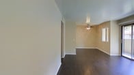 This single-floor space presents a straightforward and functional layout designed for comfortable everyday living. The interior is characterized by rich, dark hardwood flooring that runs throughout, providing a warm and grounding contrast to the light neutral walls. The main living area is open and airy, with smooth ceilings accented by a ceiling fan with an integrated light fixture, ensuring year-round comfort and ambient illumination. The walls are painted in warm beige and soft white tones that complement the dark flooring and contribute to an inviting atmosphere. White baseboards frame each room cleanly, adding a crisp finishing detail to the overall aesthetic. A doorway visible from the main living area leads to an adjoining room, suggesting a connected and accessible floor plan. A window allows natural light to filter into the space, brightening the interior and enhancing the open feel. The layout is clean and uncluttered, making it well-suited for a variety of furnishing arrangements. Overall, the space combines practical design with warm material choices to create a welcoming and livable environment.