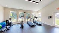 This fitness center offers a thoughtfully designed workout space that accommodates a wide range of exercise needs. The room features soaring ceilings with a recessed, framed skylight detail that adds architectural interest while contributing to the bright, open atmosphere. Recessed lighting complements the abundant natural light that pours in through a bank of large windows along one wall, framing lush outdoor views of mature trees and an adjacent pool area.

The flooring throughout is durable rubber gym matting, providing cushioning and traction suited for all types of physical activity. The cardio zone is well-appointed with multiple treadmills and an elliptical trainer, all positioned to take full advantage of the scenic outdoor views. On the opposite side of the space, a cable-based multi-function strength machine serves as the centerpiece of the strength training area, complemented by a dumbbell rack with a range of free weights, medicine balls, a stability ball, and aerobic step platforms. A workout reference chart is mounted on the wall for user guidance.

Two access points — including a clearly marked exit door — provide convenient entry and egress. The neutral, light-toned walls and clean layout create a motivating yet calm environment, making this fitness center a functional and inviting space for daily exercise.