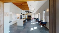This single-floor community clubhouse in Redmond, Oregon, is designed with a contemporary aesthetic that seamlessly blends warm natural materials with clean, modern finishes. The space is defined by dramatic vaulted ceilings that soar overhead, punctuated by a striking wood slat ceiling detail above the kitchen zone and a distinctive timber-framed architectural element anchoring the central media area.

The open-concept layout flows from a fully equipped kitchen featuring a large island with a wood-accented base, white quartz countertops, subway tile backsplash, stainless steel appliances, and pendant lighting, into a spacious lounge area furnished with a leather sectional sofa and a large flat-screen television set within a custom wood surround.

A dedicated seating area runs along a wall of tall, narrow windows that flood the space with natural light, outfitted with mid-century modern-style chairs and small café tables, creating an inviting social atmosphere. At the far end, a billiards table anchors a game area set against a teal accent wall with a built-in counter and bar stools. Large-format slate tile flooring unifies the entire space, contributing to the polished, cohesive character of this well-appointed communal facility.