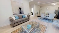 This thoughtfully designed single-floor residence exudes a calm, sophisticated ambiance through its cohesive color palette and refined furnishings. The open-concept layout seamlessly connects the living and dining areas, creating a generous, airy feel enhanced by light hardwood flooring that runs throughout the space.

The living area serves as the centerpiece of the home, anchored by a tufted gray sofa adorned with turquoise and white accent pillows. A glass-top coffee table with a metallic frame rests atop a richly patterned area rug, adding warmth and visual interest to the space. A large-scale abstract artwork on the wall provides a striking focal point, lending an artistic character to the room.

The dining area is visible just beyond the living space, featuring upholstered seating arranged around a round blue area rug, continuing the home's soft blue accent theme. Recessed lighting throughout the ceiling provides clean, even illumination, complementing the bright, white walls that keep the interior feeling open and fresh.

The transition between rooms is smooth and intuitive, with well-proportioned doorways and clear sightlines that enhance the sense of flow. Overall, the space strikes a balance between comfort and elegance, making it a welcoming and visually cohesive environment.
