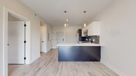 This contemporary single-floor residence is thoughtfully designed with a clean, modern aesthetic that balances style and functionality. The open-concept layout centers around a striking kitchen, where a large navy blue island with a crisp white countertop serves as the focal point. Twin pendant lights hang above the island, adding a refined touch to the space. The kitchen is fully equipped with stainless steel appliances, including a French door refrigerator, electric range, and over-the-range microwave. A dramatic dark marble-style backsplash contrasts beautifully against the combination of white upper cabinets and dark lower cabinetry, creating visual depth and sophistication.

Light-toned hardwood-style flooring runs seamlessly throughout the unit, lending warmth and continuity to each room. The bedroom is a bright, tranquil space featuring a large window that invites natural light and offers views of the surrounding exterior. Neutral wall tones and clean trim work keep the room feeling airy and versatile.

The bathroom is finished with dark veined marble-style floor tiles, a soaking tub, and an elegant white marble-patterned surround, giving the space a spa-like quality. Throughout the unit, the design reflects a cohesive modern palette with high-quality finishes that are both visually appealing and practical.