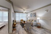 This single-story residence offers a thoughtfully designed layout encompassing two bedrooms, two bathrooms, a kitchen, and a hallway. The living area serves as the heart of the home, featuring wood-style flooring, a ceiling fan, and a large window dressed with sheer curtains that flood the space with natural light. The room comfortably accommodates a full seating arrangement, creating a welcoming and functional gathering space.

The kitchen is efficiently laid out with light wood cabinetry, granite-style countertops, a double-basin sink, a dishwasher, and white appliances including a range and refrigerator. A pass-through counter connects the kitchen to the living area, enhancing the open feel of the space. Light tile flooring and recessed lighting complete the kitchen's clean, practical design.

Both bedrooms feature plush carpeting, ceiling fans, and large windows with sheer curtains that invite natural light while maintaining privacy. The primary bedroom offers a notably spacious feel, while the second bedroom is equally bright and comfortable.

The two bathrooms are finished with warm wood vanities, stone-style countertops, and full tub-shower combinations with white tile surrounds. Light flooring and clean, neutral walls give both bathrooms a bright, functional character. A connecting hallway ties all rooms together in a cohesive and easy-to-navigate floor plan.