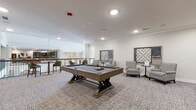 Located at 9246 Highland Creek Pkwy in Charlotte, NC, this single-floor community game and recreation room is thoughtfully designed to encourage social interaction and leisure. The space features neutral-toned, textured carpeting throughout, crisp white walls accented with crown molding, and recessed lighting that keeps the interior bright and welcoming.

A centerpiece pool table with a dark felt surface and a distinctive trestle-style base occupies the main area, complemented by a wall-mounted cue rack nearby. Adjacent to the pool area, a full-length shuffleboard table is set against a sage green accent wall, which also features a large flat-screen television and decorative artwork, creating an entertainment-focused nook.

A dedicated lounge corner showcases oversized, interactive wall-mounted Scrabble boards alongside patterned armchairs and small accent tables, offering a comfortable setting for games and conversation. Additional high-top seating with bar-height chairs is positioned along a railing, providing views into a neighboring area. The overall ambiance is modern yet relaxed, combining purposeful recreation amenities with inviting residential décor, making it a well-rounded gathering space for residents to enjoy.