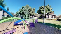 This outdoor common area serves as a central gathering space within a residential community, offering a variety of amenities designed for both recreation and relaxation. The space is anchored by a vibrant children's playground equipped with a multi-colored play structure featuring climbing elements, a slide, swings, and stepping stairs, all finished in bright green, blue, and purple. The playground is set atop a wood chip surface, providing a safe and cushioned ground cover for young users.

Surrounding the play area, expansive stretches of well-kept green lawn offer open space for outdoor activities. Mature, full-canopy trees are dispersed throughout the grounds, casting generous shade and contributing to a pleasant, park-like atmosphere. Raised mulched planting beds frame sections of the lawn, adding visual interest and low-maintenance landscaping detail.

Covered carport parking structures are visible along the perimeter, providing residents with protected vehicle storage. Additional site lighting via tall poles ensures the common area remains functional during evening hours. A community information sign is posted near the playground, offering residents relevant guidelines for the space. Overall, the grounds present a clean, well-organized outdoor environment suited to everyday community life.