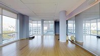 This single-floor fitness studio offers a spacious, open-concept layout designed to accommodate a variety of exercise disciplines, from yoga and Pilates to dance and group training. The room features wide-plank hardwood flooring throughout, providing a warm, resilient surface well-suited for active use. Floor-to-ceiling windows line multiple walls, flooding the interior with abundant natural light while framing expansive urban views of the surrounding cityscape. The generous glazing creates an airy, energizing atmosphere that enhances the overall workout environment. Cylindrical structural columns are positioned thoughtfully within the space, adding an architectural element without significantly interrupting the open floor plan. A mirrored wall on one side serves as a functional fitness feature, visually expanding the room while allowing participants to monitor their form. The ceiling is finished with a clean, recessed tile panel design that contributes to the studio's polished, professional aesthetic. Exercise equipment, including stability balls and a step platform, can be seen stored within the space. The overall ambiance is modern and minimalist, with a neutral blue-grey and white color palette that promotes focus and calm throughout the studio.