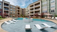 Located at 5350 Pinehurst Park Dr in Charlotte, NC, this outdoor amenity space is a thoughtfully designed resort-style retreat set within a multi-story residential community. The centerpiece is a generously sized, freeform swimming pool with crystal-clear blue water, complemented by a dedicated in-water lounge area featuring sleek, modern water loungers — ideal for relaxing while partially submerged. A separate circular spa with a central fountain jet provides a distinct relaxation zone, surrounded by manicured landscaping and curved walkways. The pool deck is finished in a warm tan concrete surface and lined with rows of contemporary chaise lounges and market umbrellas, offering ample space for sunbathing and outdoor lounging. Lush greenery, ornamental Japanese maple trees with vivid red foliage, and neatly trimmed shrubs add natural color and texture throughout the courtyard. Outdoor dining areas with tables and umbrellas are tucked into landscaped nooks along the perimeter. The space is framed on all sides by the multi-story building facade, which features a mix of brick, siding, and large windows with private balconies overlooking the courtyard. Classic lamp post lighting adds charm and ensures the space is well-lit in the evening hours.