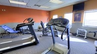 This fitness center offers a dedicated space for cardiovascular and general exercise, thoughtfully designed to support an active lifestyle. The room is finished with warm, energizing orange accent walls complemented by a neutral lower half, creating a motivating atmosphere for workouts. A drop ceiling with recessed lighting provides consistent, even illumination throughout the space, while windows allow natural light to brighten the room. The flooring is covered in durable, patterned carpet suited for a fitness environment. The center is equipped with multiple treadmills and elliptical cross-trainers, offering a range of cardio options for users of varying fitness levels. The machines are arranged to allow comfortable spacing and ease of movement between equipment. A wall-mounted television is positioned to keep users entertained during their workouts. Additional fitness accessories and equipment are visible along the perimeter of the room, providing further options for strength and conditioning exercises. The overall layout is practical and functional, making efficient use of the available floor space while maintaining an open, accessible feel throughout the fitness area.