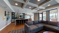 This thoughtfully designed single-floor space showcases an open-concept layout that seamlessly connects a living area, kitchen bar, and wet bar into one cohesive, elegant environment. The architectural centerpiece is the striking coffered ceiling with crisp white molding, which adds depth and a refined character throughout the main living and kitchen zones.

The living area is anchored by a generous sectional sofa in a textured grey fabric, set atop a plaid area rug over warm hardwood flooring. A wood-framed fireplace with a stone surround serves as a focal point, topped by a large wall-mounted television, creating a comfortable yet polished atmosphere. Sheer floor-to-ceiling curtains frame large sliding glass doors that open to an outdoor terrace, flooding the interior with natural light and providing views of the exterior patio space.

The kitchen bar features a dramatic waterfall-edge marble island with bar seating, illuminated by a pair of pendant lights with an industrial-modern aesthetic. Adjacent to the island, a dedicated wet bar runs along one wall, finished in deep forest green cabinetry with glass-front upper cabinets, dark countertops, a sink, and integrated appliances. Curated framed artwork throughout adds personality and a gallery-like quality to the overall space.
