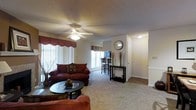 This single-floor residence offers a thoughtfully designed open-concept layout that creates a comfortable and functional living environment. The living area serves as the heart of the home, featuring plush wall-to-wall carpeting that adds warmth underfoot, and a ceiling fan with integrated lighting that ensures year-round comfort and ambient illumination. Large windows dressed with decorative valances and vertical blinds allow generous natural light to fill the space while maintaining privacy.

The living area flows seamlessly into a dining and kitchen space, visible through the open floor plan. The kitchen and dining zone features hard flooring, a raised bar-height counter with seating, and additional natural light from a second window — creating a bright and inviting area for meals and gatherings.

A hallway connects the main living space to additional rooms, with smooth transitions between carpet and hard flooring throughout. The walls are finished in neutral tones, complemented by a textured ceiling, giving the space a clean and classic aesthetic. White trim and door casings add a crisp, polished detail throughout. Overall, the residence offers a practical and welcoming single-level layout suited for comfortable everyday living.