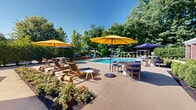 Located at 7 Archstone Ave in Tewksbury, Massachusetts, this outdoor amenity space centers around a generously sized rectangular swimming pool with crystal-clear turquoise water. The pool features wide entry steps on multiple sides, making it easily accessible, and is enclosed within a well-maintained fenced perimeter for safety and privacy.

Surrounding the pool is an expansive pool deck finished in light-toned concrete and warm wooden decking, offering ample space for lounging and outdoor entertaining. Colorful market umbrellas — in shades of yellow and purple — shade clusters of tables and chairs, providing comfortable spots to relax in the sun. Cushioned lounge chairs and outdoor seating arrangements are thoughtfully positioned throughout the deck area.

A covered pavilion anchors one side of the space, featuring a classic pitched roof, brick columns, and large windows that invite natural light. This structure provides a shaded retreat and additional seating for residents and guests. Lush, mature trees form a natural green backdrop along the perimeter, creating a sense of seclusion and tranquility. Neatly trimmed hedges and decorative plantings further enhance the landscaped setting.

Together, these elements create a resort-style outdoor environment that is both functional and inviting, ideal for warm-weather recreation and relaxation.