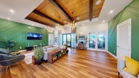 This single-story community clubhouse delivers a thoughtfully designed gathering space that blends modern style with warm, natural materials. The open-plan lounge is immediately distinguished by its vaulted wood-planked ceiling, accented by dark exposed beams that add architectural depth and a resort-inspired character. Two dramatic sputnik-style chandeliers serve as focal points, casting ambient light across the expansive hardwood flooring that runs throughout the space.

The lounge area is anchored by a comfortable seating arrangement featuring a neutral sofa with colorful accent pillows, a low-profile coffee table, and a pair of deep green velvet accent chairs. A bold, geometrically patterned green accent wall—complete with diagonal trim detailing and a wall-mounted television—adds vibrant color and visual interest to one end of the room.

Natural light floods the interior through multiple large windows and glass French doors, which open toward an outdoor area with views of lush landscaping and a pool. Sheer curtain panels frame the doors, softening the transition between indoors and out. A dedicated coffee station and a glass-enclosed room are visible, suggesting additional amenity spaces. Decorative indoor plants and potted grasses enhance the fresh, tropical ambiance throughout.