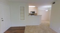 This single-story space showcases an open-concept layout that connects the living area to the kitchen through a curved peninsula-style counter with a granite or stone-look surface. The kitchen is equipped with white cabinetry, stainless steel appliances including a built-in microwave, and overhead lighting that brightens the cooking area. A practical pass-through design allows for easy interaction between the kitchen and the adjoining living space.

The living area features a combination of wood-look hard flooring and neutral-toned carpeting, creating a warm and welcoming atmosphere. A built-in shelving unit is recessed into one wall, offering display or storage space while adding architectural interest to the room. The walls are finished in a soft, neutral tone throughout, contributing to a clean and cohesive aesthetic. A pendant light fixture is partially visible, adding a decorative element to the space. The overall design is functional and straightforward, with an emphasis on open sightlines and practical built-in features.