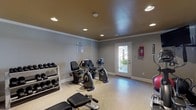 Located at 1 Hampton Pl in Birmingham, Alabama, this single-floor fitness center is a well-appointed exercise facility designed to support a comprehensive workout routine. The room features neutral gray walls accented by crisp white crown molding and baseboards, with a warm-toned ceiling that lends a distinct, polished character to the space. Recessed lighting is distributed evenly throughout, ensuring bright, consistent illumination across the entire room.

The facility is equipped with a diverse selection of cardio machines, including multiple upright and recumbent stationary bikes and an elliptical trainer, accommodating a range of fitness preferences. A commercial-grade Hoist cable machine serves as the centerpiece for strength training, complemented by a cushioned workout bench and a well-stocked rack of dumbbells in varied weights. A stability ball and additional accessories round out the available equipment.

The flooring transitions between durable speckled epoxy-style material in the cardio area and carpet near the strength equipment, providing both functionality and comfort. A row of small horizontal windows near the ceiling on one wall allows natural light to filter in, while a glass-paneled exterior door opens to an outdoor area, enhancing ventilation and connectivity to the surrounding environment. A mounted flat-screen television and posted fitness rules complete this practical, community-oriented workout space.