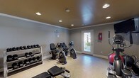 Located at 1 Hampton Pl in Birmingham, Alabama, this single-floor fitness center is a well-appointed exercise facility designed to support a comprehensive workout routine. The room features neutral gray walls accented by crisp white crown molding and baseboards, with a warm-toned ceiling that lends a distinct, polished character to the space. Recessed lighting is distributed evenly throughout, ensuring bright, consistent illumination across the entire room.

The facility is equipped with a diverse selection of cardio machines, including multiple upright and recumbent stationary bikes and an elliptical trainer, accommodating a range of fitness preferences. A commercial-grade Hoist cable machine serves as the centerpiece for strength training, complemented by a cushioned workout bench and a well-stocked rack of dumbbells in varied weights. A stability ball and additional accessories round out the available equipment.

The flooring transitions between durable speckled epoxy-style material in the cardio area and carpet near the strength equipment, providing both functionality and comfort. A row of small horizontal windows near the ceiling on one wall allows natural light to filter in, while a glass-paneled exterior door opens to an outdoor area, enhancing ventilation and connectivity to the surrounding environment. A mounted flat-screen television and posted fitness rules complete this practical, community-oriented workout space.