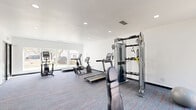 This fitness center at 6350–6400 Oakley Rd in Union City, GA offers a thoughtfully designed exercise space spread across a single floor. The room is finished with clean white walls and recessed lighting throughout, creating a bright and energizing atmosphere. A patterned carpet with bold accent lines covers the floor, adding a modern aesthetic while providing comfort underfoot.

The space is well-appointed with a comprehensive selection of exercise equipment. Cardio options include multiple Matrix treadmills and a stationary bike, positioned near large windows that allow natural light to fill the room and offer views of the surrounding greenery. Strength-training equipment includes a Matrix functional trainer cable machine, a multi-function selectorized weight machine for leg and upper body exercises, a dedicated weight bench, and a full dumbbell rack with a range of free weights. A stability ball rounds out the functional training offerings.

The layout is open and spacious, allowing for comfortable movement between stations. Two interior doors provide access to additional areas, and a glass-panel entry door connects the space to the exterior. The overall ambiance is clean, modern, and functional, making this fitness center a practical and inviting facility for residents and users seeking a well-rounded workout experience.