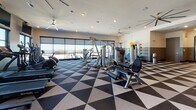 This modern fitness center is designed to provide a comprehensive workout experience within a thoughtfully organized, single-floor layout. The space is immediately distinguished by its bold geometric flooring — a striking black, white, and gray triangular tile pattern that adds a dynamic visual energy throughout.

Large floor-to-ceiling windows line the walls, flooding the interior with abundant natural light and offering expansive views of the surrounding outdoor environment, creating an open and motivating atmosphere. Ceiling fans are positioned throughout, ensuring comfortable air circulation during workouts.

The cardio zone features a row of SportsArt treadmills and elliptical machines positioned directly in front of wide windows, complemented by wall-mounted flat-screen televisions for entertainment. Nearby, a selection of rowing machines and additional cardio equipment rounds out the aerobic offerings.

The strength training area is well-appointed with a full rack of dumbbells, weight benches, and cable machines, supported by floor-to-ceiling mirrors that enhance the sense of space. A dedicated open-floor stretching and functional training zone provides room for mat work and flexibility exercises. Built-in cubby storage and informational signage near the entrance add practical convenience. Recessed lighting throughout ensures the space is bright and welcoming at all hours.
