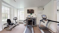 This fitness center occupies a single-floor space divided into two connected rooms, blending classic architectural character with modern exercise equipment. The first room is a standout, featuring a decorative marble fireplace as a focal point, above which a flat-screen television is mounted for entertainment during workouts. Two treadmills and an upright stationary bike are positioned throughout, while a wall-mounted split-system air conditioning unit ensures a comfortable environment. Natural light filters in through traditional sash windows, complementing the crisp white walls, crown molding, and neutral carpet flooring. A coat rack near the door adds a practical touch. The second room continues the clean, bright aesthetic with additional large sash windows and track lighting overhead. This area houses an elliptical trainer, a recumbent bike, and a multi-function strength machine, alongside an adjustable bench and a full dumbbell rack arranged in front of a large wall mirror. A second flat-screen television is mounted on the wall for added convenience. Together, the two rooms provide a comprehensive workout environment that balances functional fitness amenities with the refined character of the building's traditional architecture.