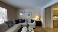 This single-floor space centers around a well-appointed living area designed for comfort and functionality. The living room features a generous sectional sofa in a neutral cream tone, accented with navy and patterned throw pillows that add visual interest and a pop of color. A textured carpet grounds the seating area, contributing to the room's warm, relaxed feel.

The walls are finished in a soft beige tone, complemented by a textured ceiling that adds subtle dimension overhead. Decorative framed artwork with geometric patterns adorns the walls, reinforcing a cohesive, modern-casual aesthetic throughout the space.

A dark wood side table positioned in the corner holds a classic table lamp, casting a warm, ambient glow that enhances the room's cozy atmosphere. Greenery in the form of potted plants adds a natural, refreshing element to the décor. A mirrored tray coffee table accessory and a patterned ottoman further contribute to the layered, thoughtfully styled interior. Natural light appears to supplement the interior lighting, keeping the space feeling bright and welcoming overall.