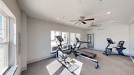 This single-floor fitness center at 10742 Freehill Street in Houston, Texas, offers a clean, modern workout environment designed to support a comprehensive cardio routine. The space is finished with neutral gray walls and low-pile carpet flooring, creating a practical and comfortable exercise environment.

The room is generously appointed with a variety of cardio equipment, including treadmills, an elliptical trainer, recumbent stationary bikes, and a rowing machine, providing users with diverse training options. A full-length mirrored wall spans one side of the room, enhancing the sense of openness and allowing users to monitor their form during workouts.

Natural light fills the space through multiple large windows positioned along the walls, several of which frame views of an outdoor pool area, adding a pleasant visual connection to the exterior. Ceiling fans are mounted throughout to assist with air circulation, complementing the HVAC system for year-round comfort.

Recessed lighting supplements the natural light, ensuring the space remains bright regardless of the time of day. A glass-panel entry door provides easy access to and from the facility. The overall ambiance is bright, open, and functional — well-suited for a community fitness amenity focused on accessibility and everyday use.