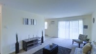 This single-floor living space offers an open and airy layout centered around a generously sized living area. The room is finished with wall-to-wall carpeting that adds warmth underfoot, complemented by clean white walls that enhance the bright, neutral atmosphere throughout.

A prominent sliding glass door dressed with vertical blinds serves as the focal point of the space, allowing abundant natural light to fill the interior while providing access to the outdoors. An additional window fitted with a wall-mounted air conditioning unit offers supplemental ventilation and climate control. A baseboard heater along the exterior wall provides further comfort year-round.

The living area comfortably accommodates key furnishings, including a low-profile media console, a tufted ottoman coffee table, and a layered area rug that defines the seating zone. A decorative wall canvas and a tall floor vase add simple artistic touches to the otherwise minimalist aesthetic.

The open-plan design allows the living area to flow seamlessly into an adjoining space, making the overall floor plan feel connected and functional. The combination of natural light, neutral finishes, and practical amenities creates a comfortable and livable environment.